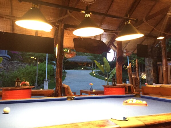 Une table de billard avec une boule, entourée de meubles en bois et illuminée par des lampes suspendues, se trouve dans une pièce dotée d’un plafond rustique et offrant une vue sur la verdure à l’extérieur.
