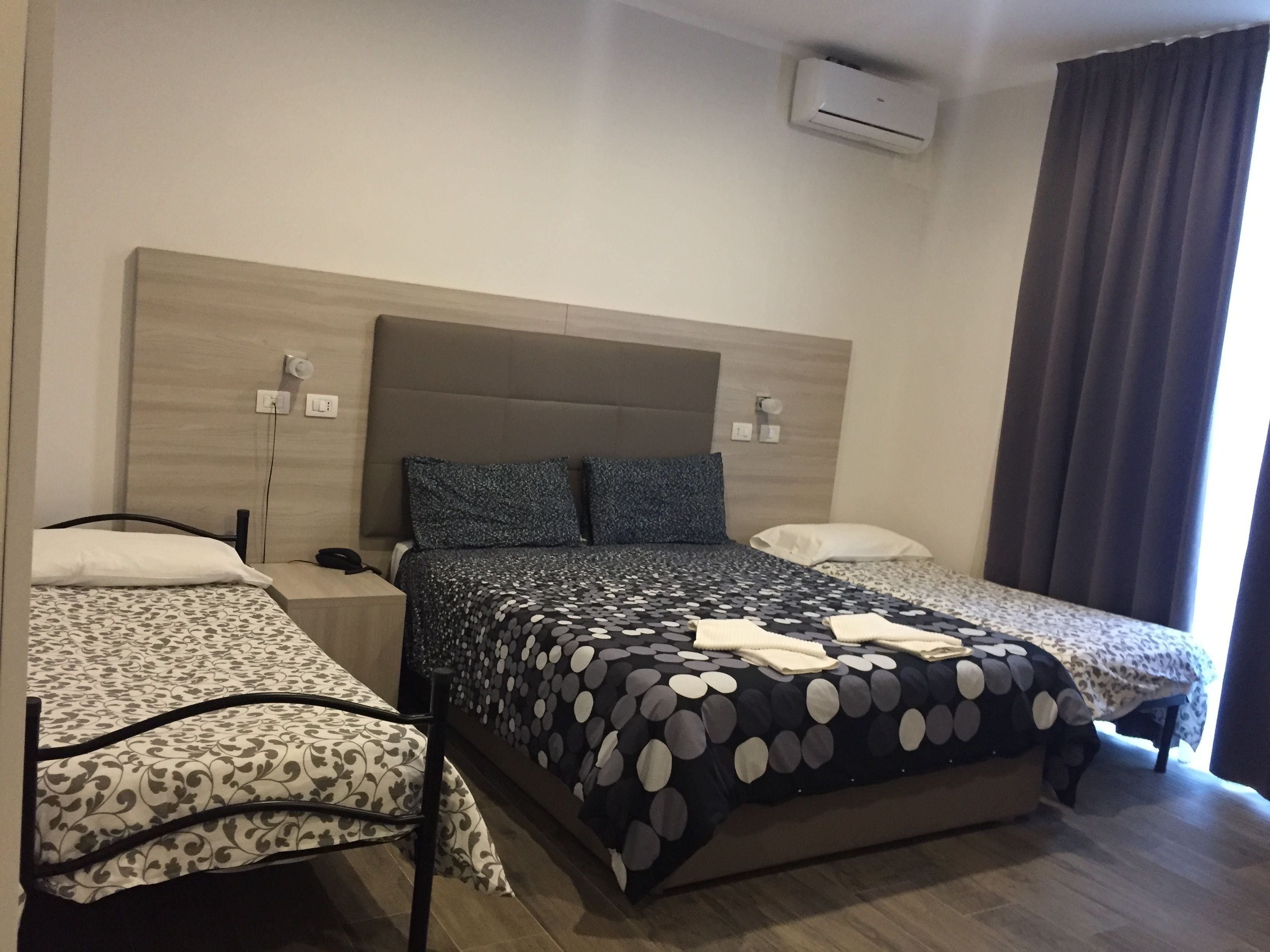 Habitación de hotel con una cama grande, dos camas más pequeñas, un cabecero, un equipo de aire acondicionado y una ventana con cortinas.