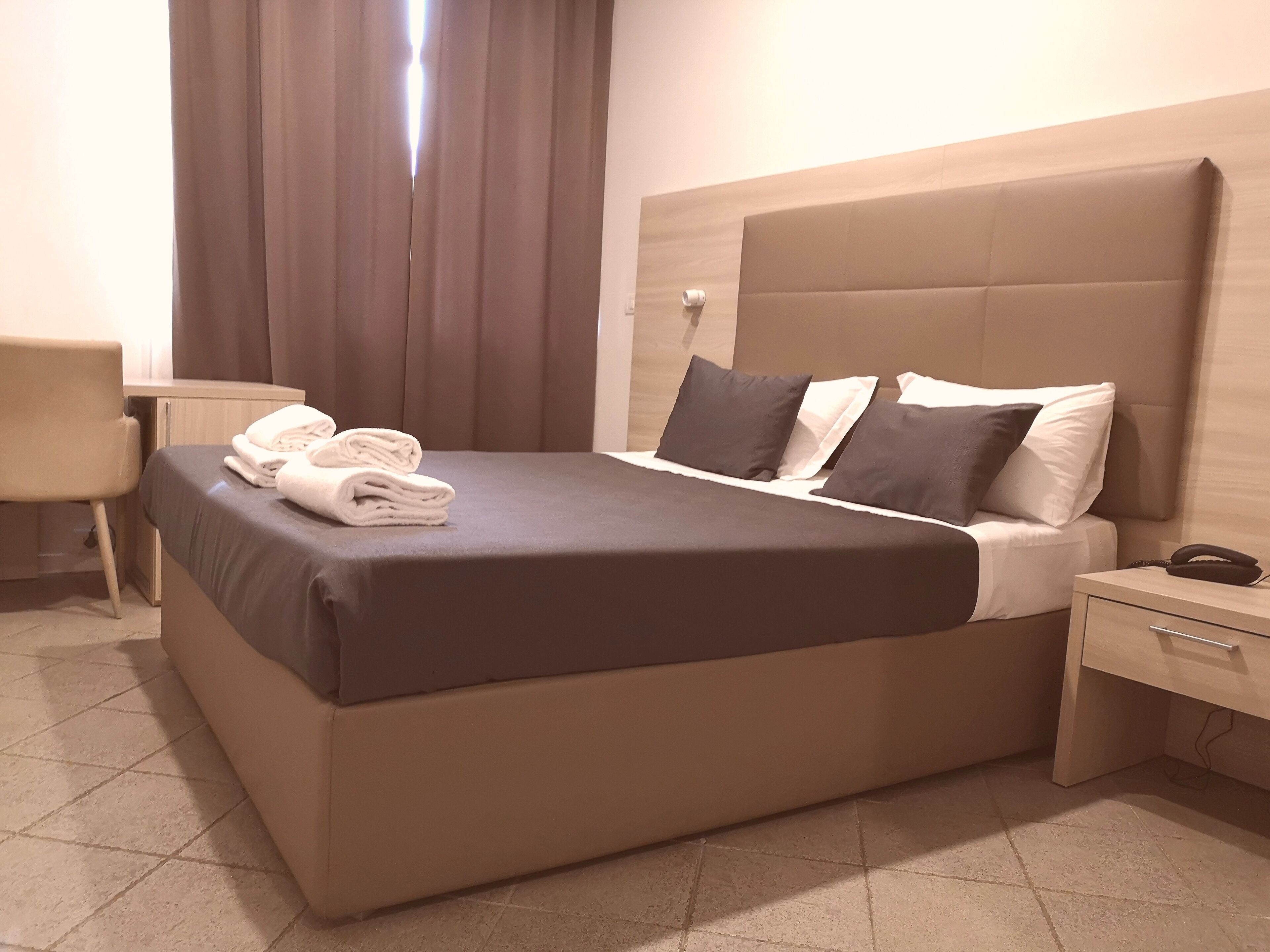 A neatly made bed with a brown headboard, a bedside table with a phone, and a chair.