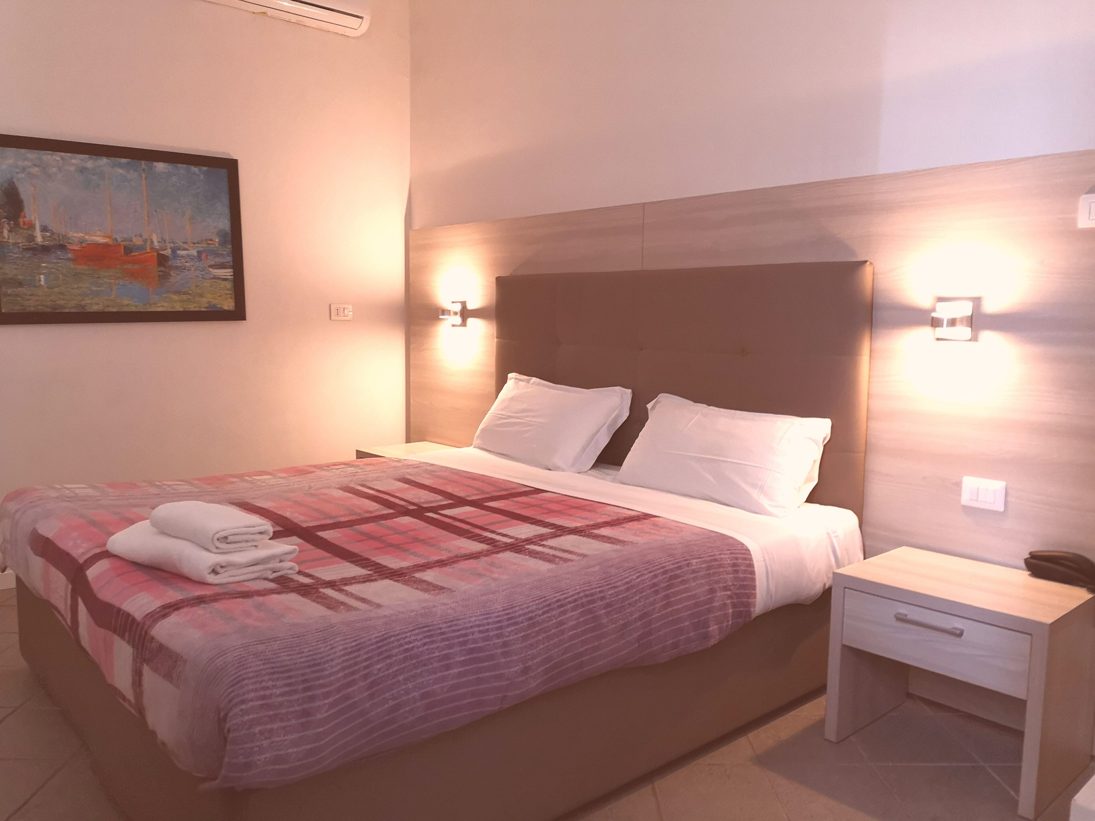 A hotel room with a bed, a nightstand, a painting on the wall, and wall-mounted lights.