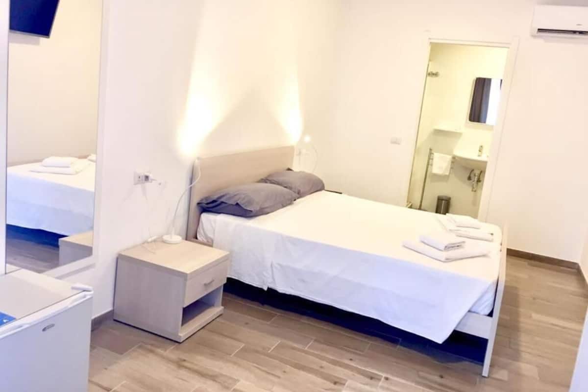 A modern hotel room with a single bed, a bedside table, a mirror, and a small desk with a chair.