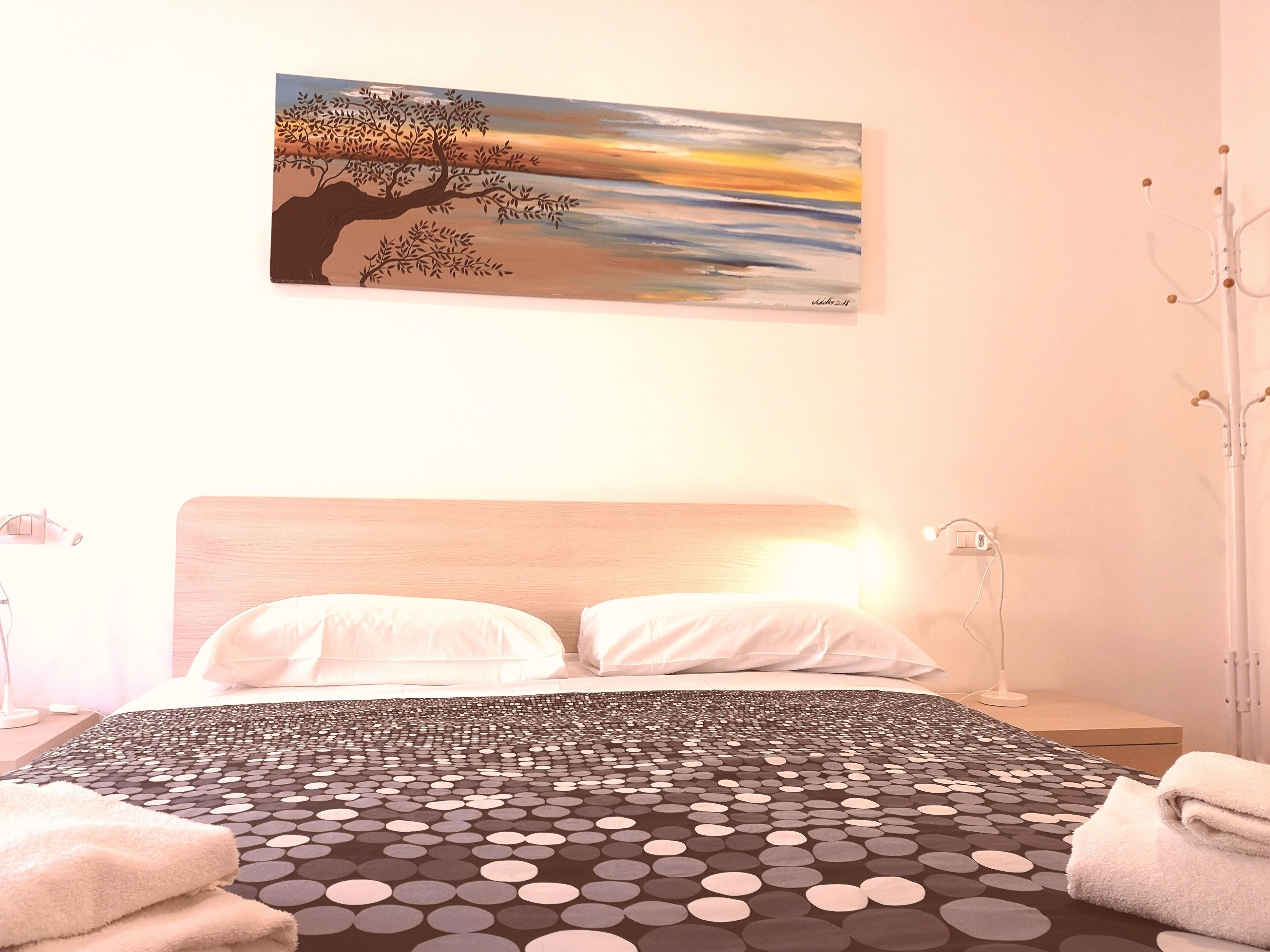 A hotel room with a bed, a painting of a tree, and a coat rack.