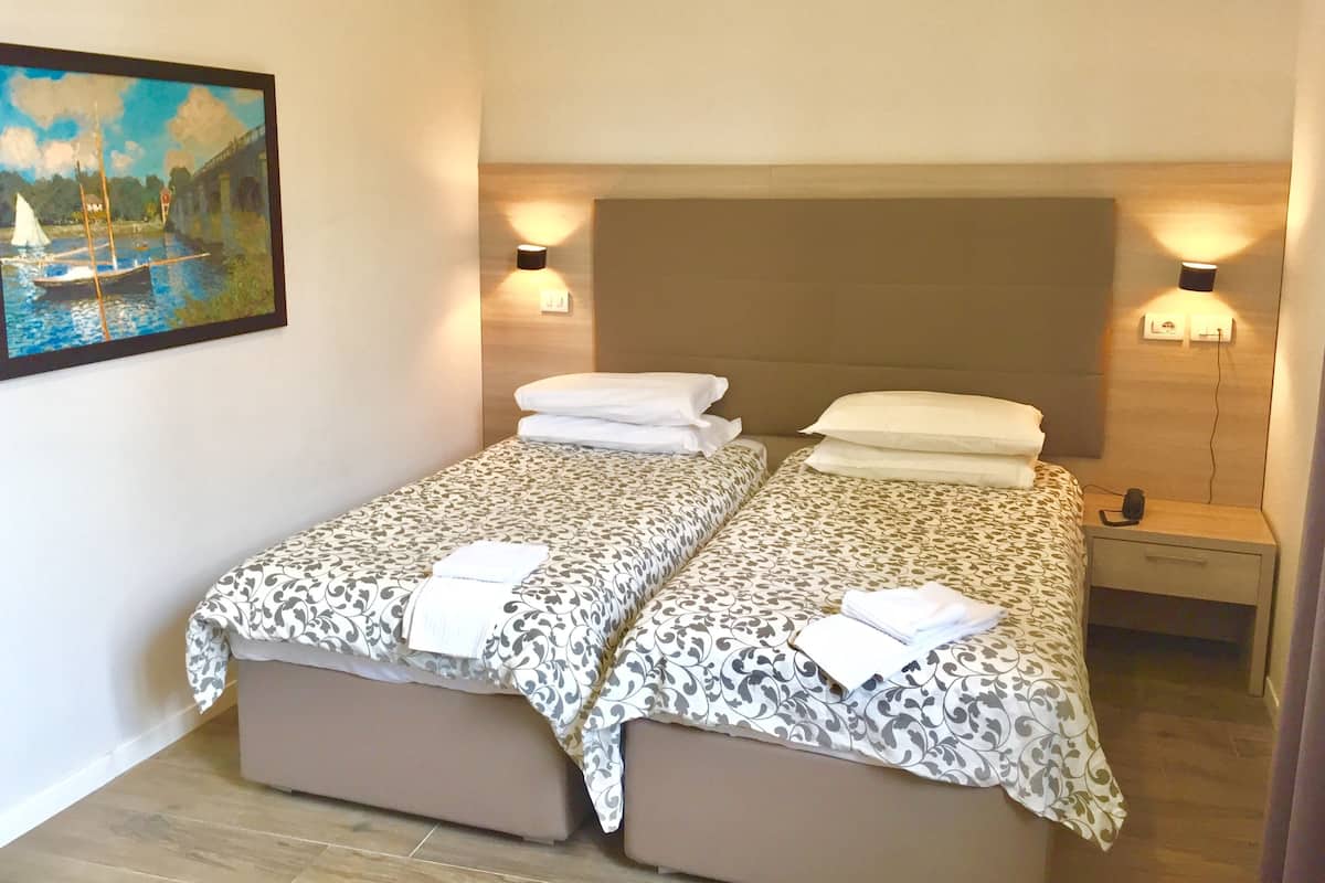A double bed with a patterned bedspread and two pillows, a bedside table with a lamp, and a painting of a river scene on the wall.