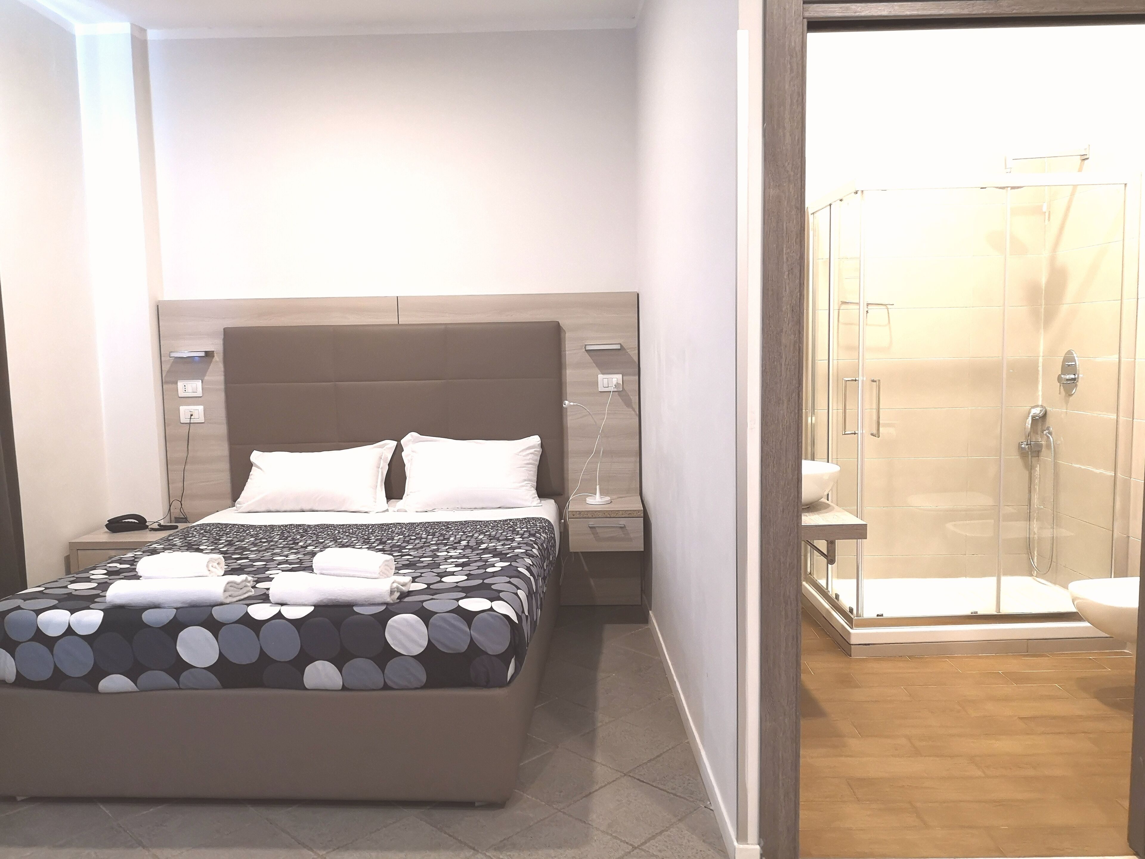 Une chambre d’hôtel moderne dotée d’un grand lit, de tables de chevet, d’une petite table et d’un espace douche séparé par une porte en verre.