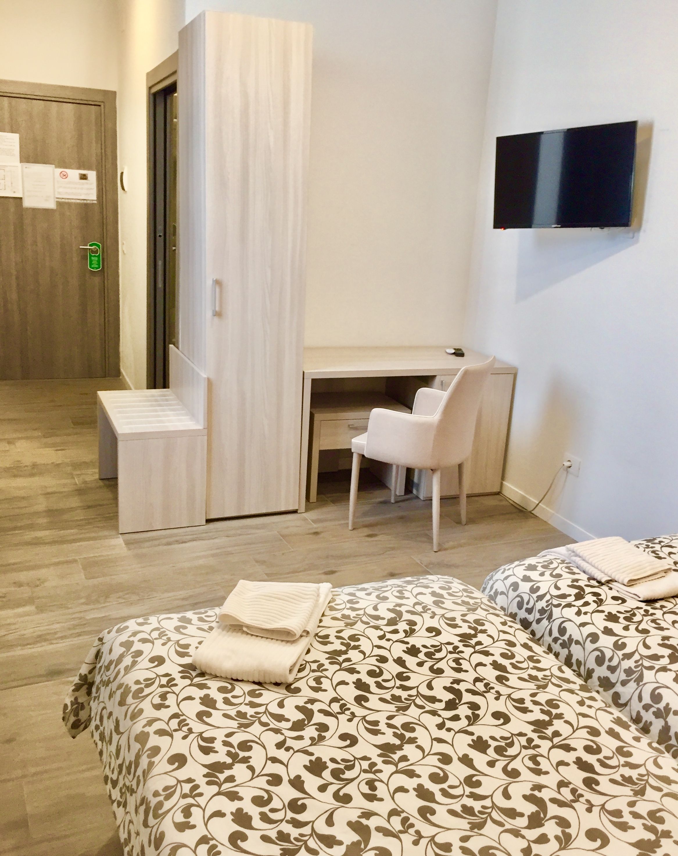 A hotel room with a bed, a desk, and a chair. There is a TV on the wall and a door with a lock.