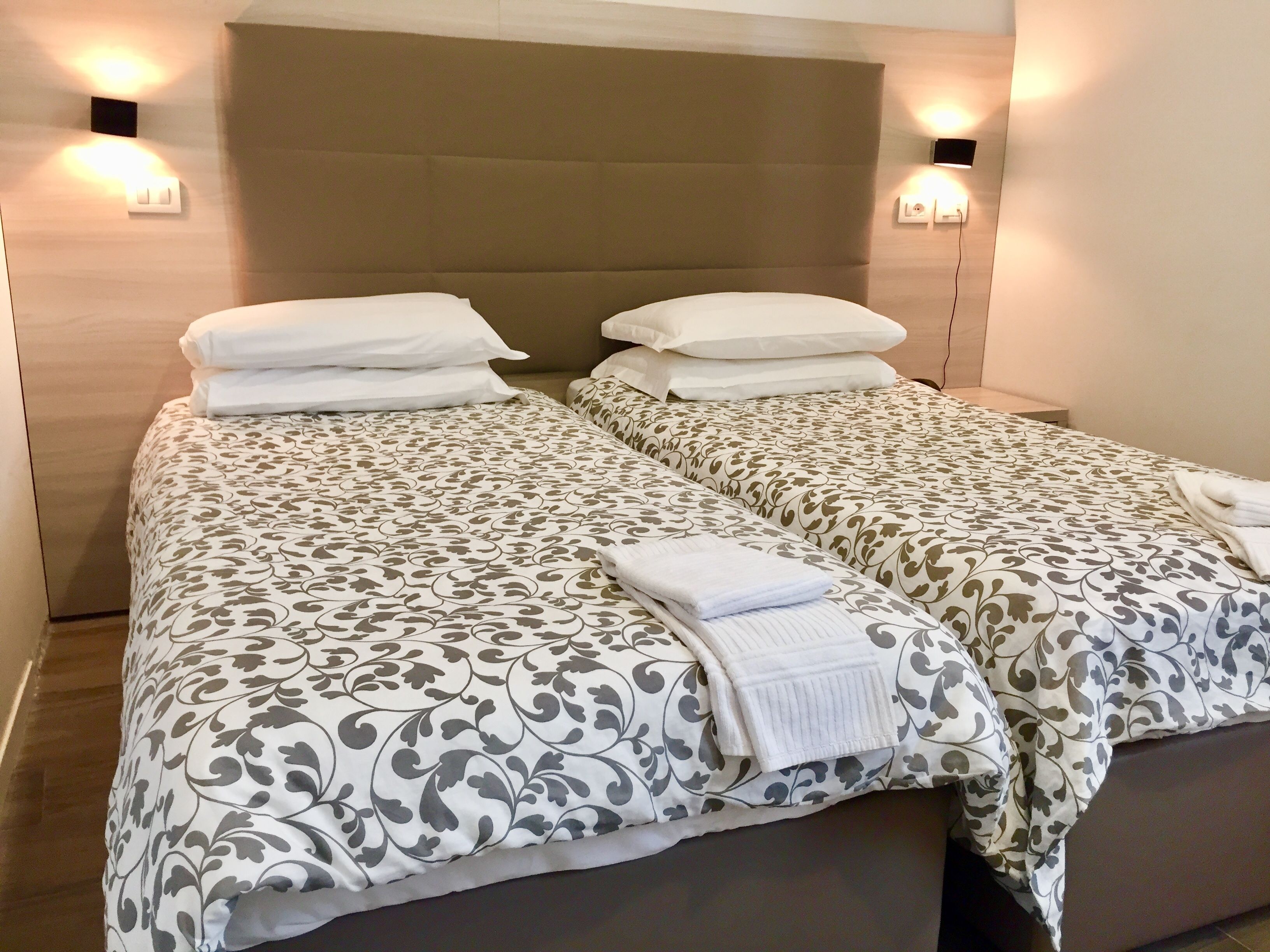 Una cama doble con una colcha estampada y dos almohadas, una mesita de noche con lámpara, y un aplique en la pared.