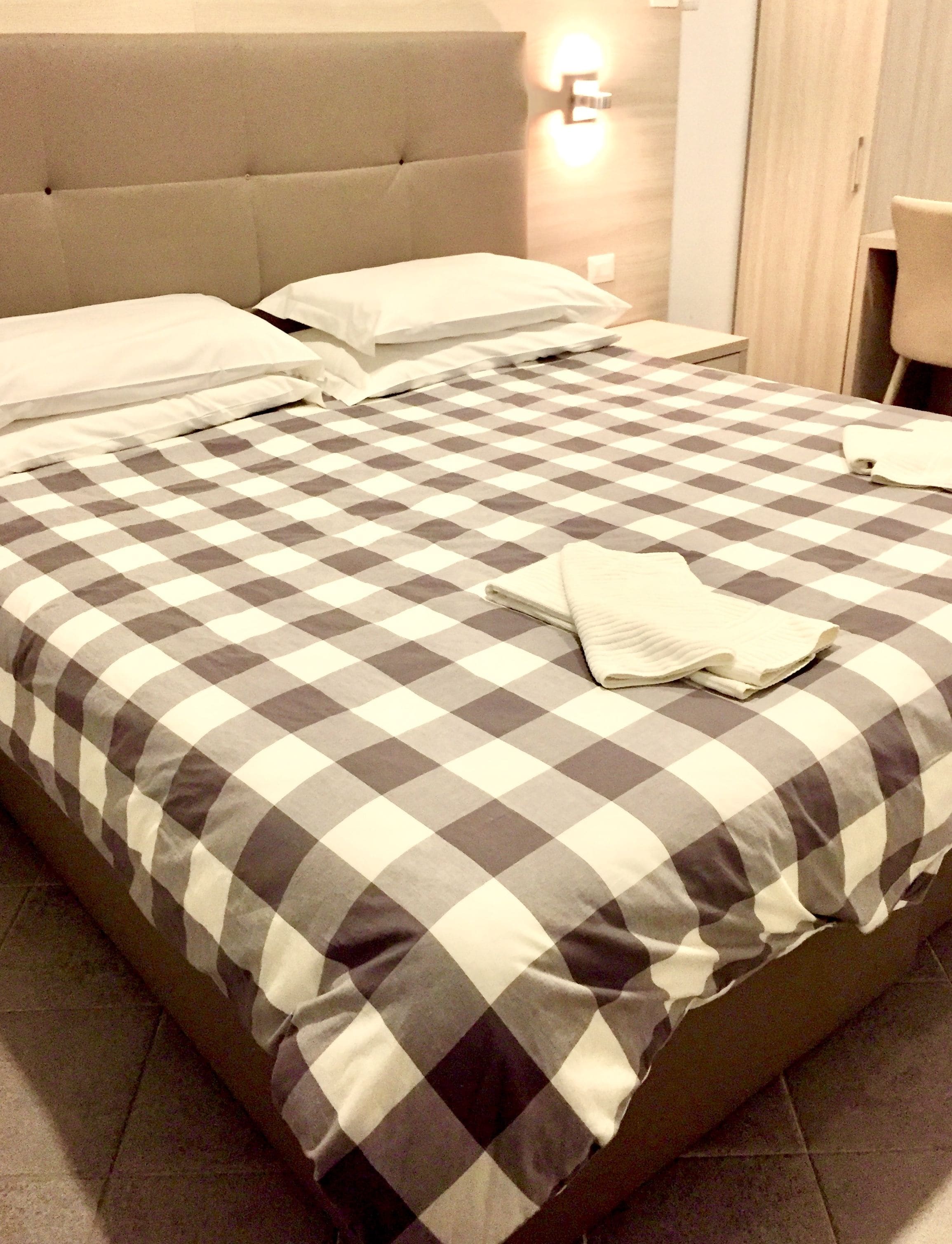 Un lit bien fait, avec une couverture à carreaux, une serviette blanche pliée et une tête de lit surmontée d’une lampe.