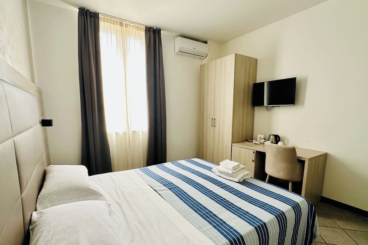A hotel room with a bed, a desk, a chair, a TV, and an air conditioning unit.