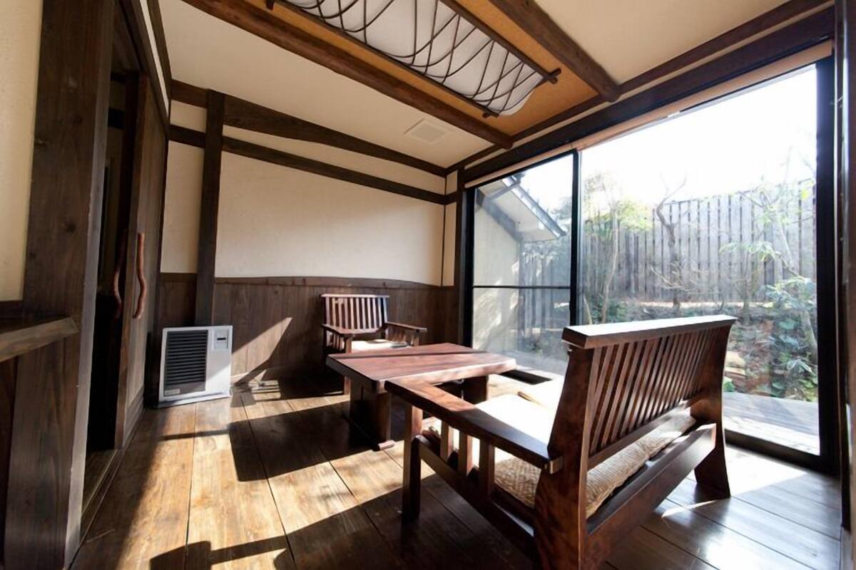 A room with a wooden floor, a table, two benches, and a large window with a view of a garden.