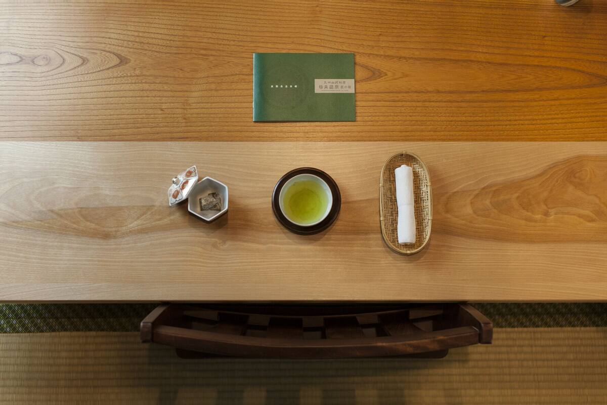 A wooden table with a green book, a small tea set, and a rolled towel.