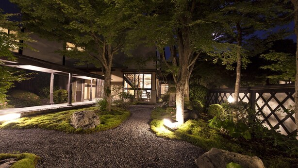 A nighttime view of a modern building with a gravel pathway, trees, and a wooden fence. - Gokuraku Onsen Takumi No Yado (Takaharu)