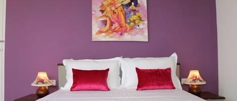 Un dormitorio con cama, dos almohadas rojas, una colcha blanca, dos mesitas de noche con lámparas y un cuadro en la pared.