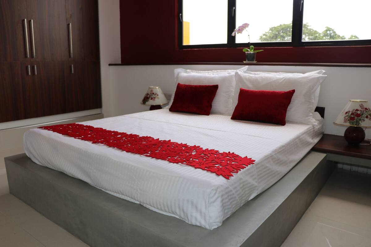 Una cama bien tendida con una colcha blanca y roja, un armario de madera y una mesita de noche con una lámpara.