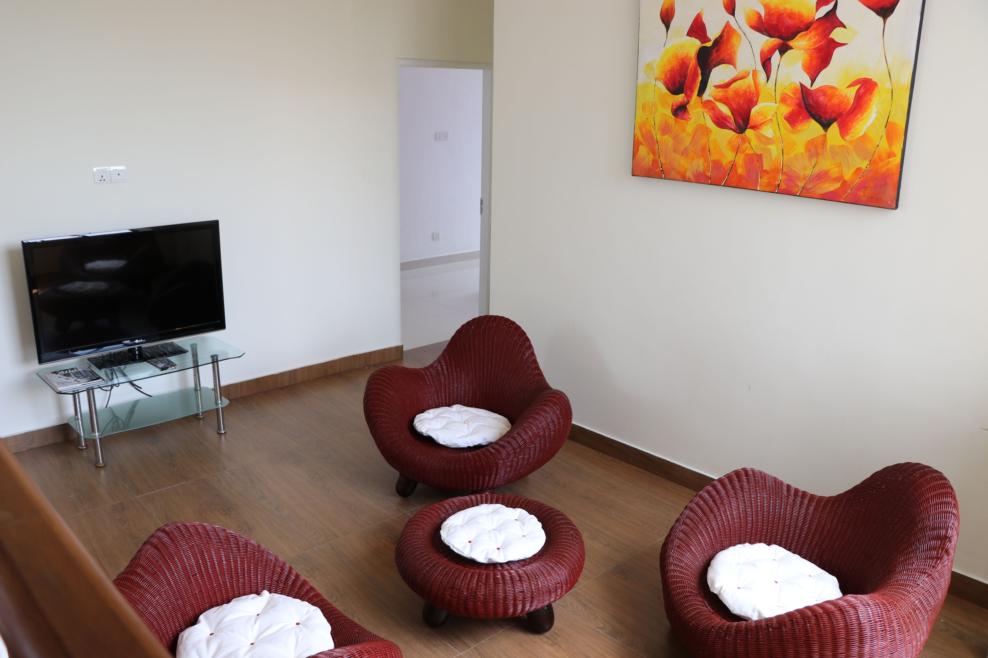 A room with a flat-screen TV on a glass table, two red wicker chairs with white cushions, and a large floral painting on the wall.
