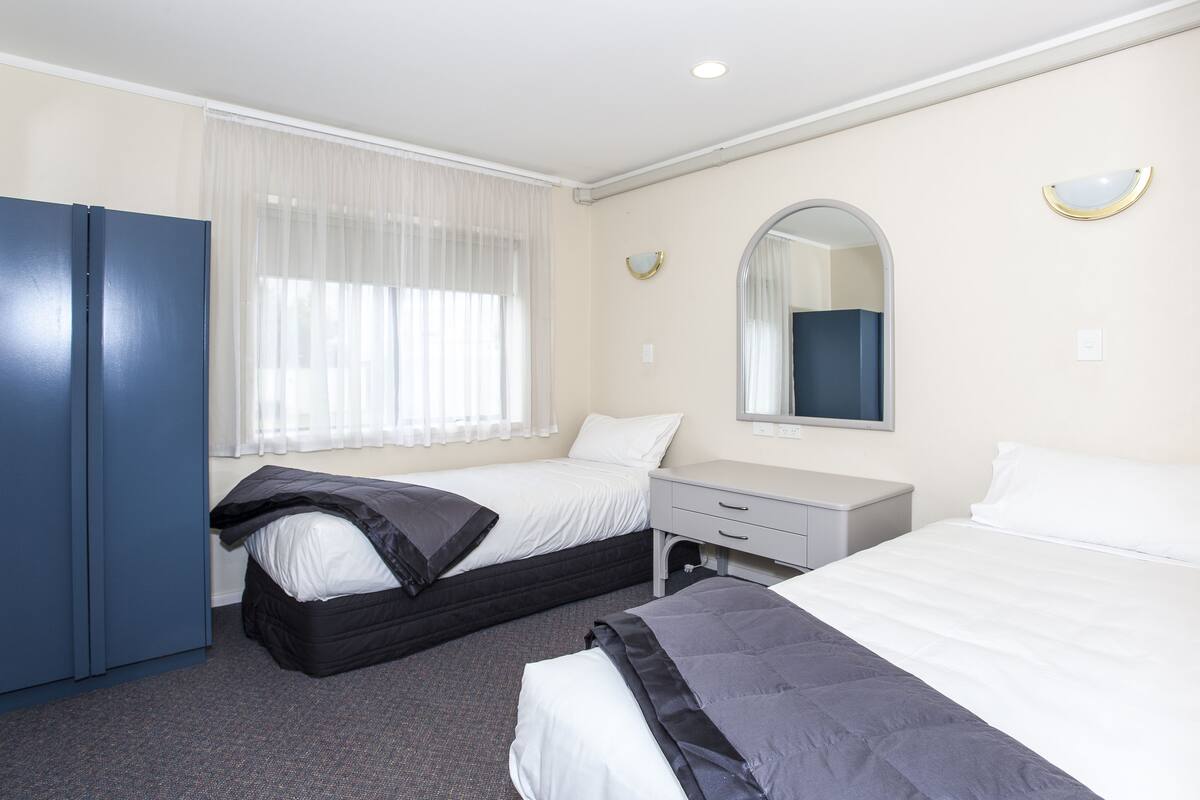 A hotel room with two beds, a wardrobe, a desk, and a window with sheer curtains.