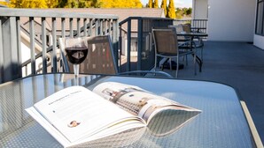 Una mesa de vidrio con una revista y una copa de vino, ubicada en un patio exterior con sillas y barandilla.