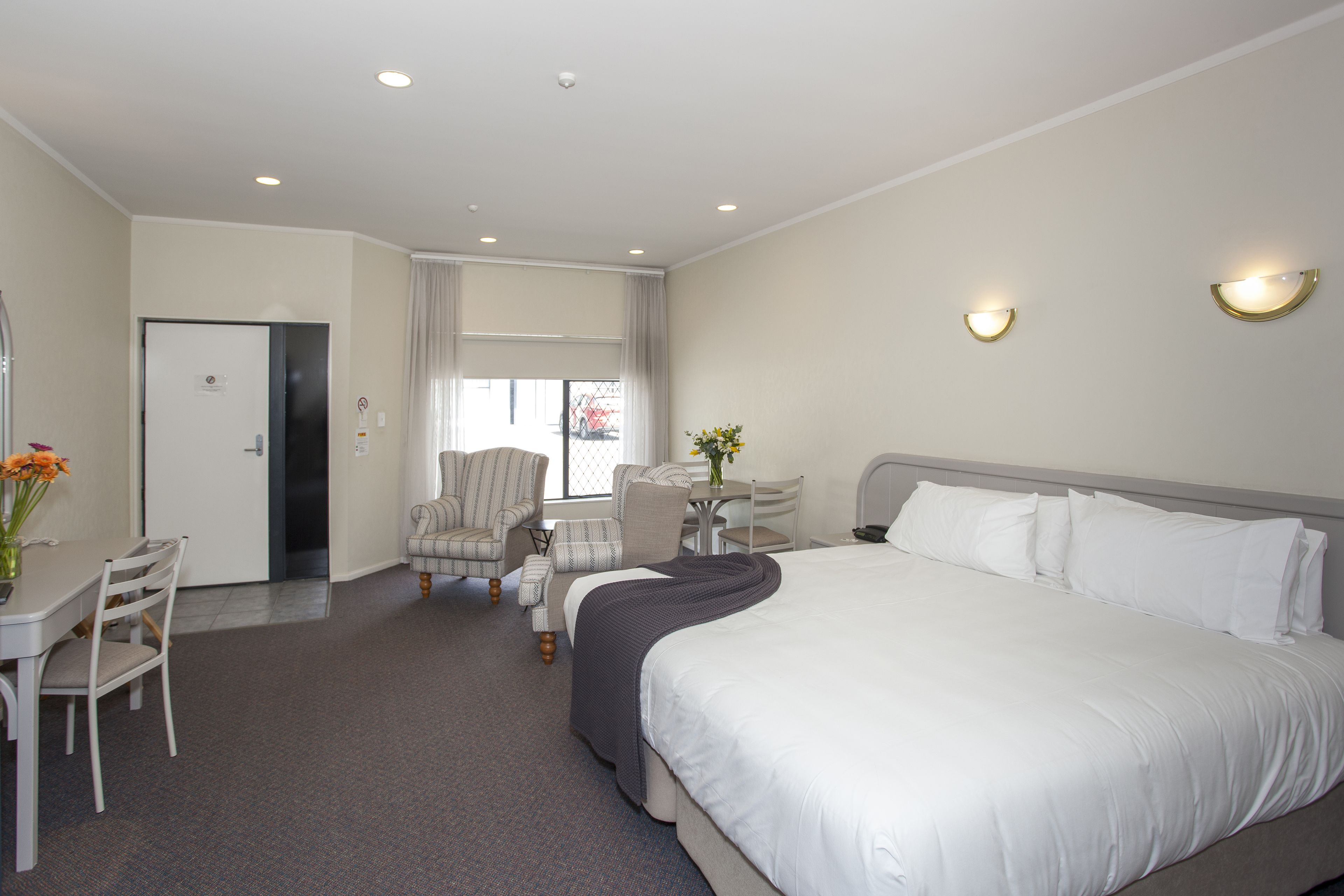 A hotel room with a large bed, a desk with a chair, a small table with a vase, and a chair.