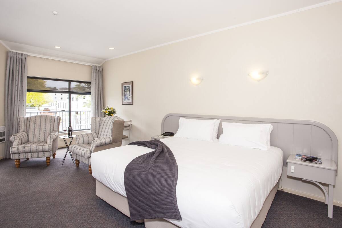 A hotel room with a large bed, two armchairs, a small table with a vase, and a nightstand with a lamp and books.