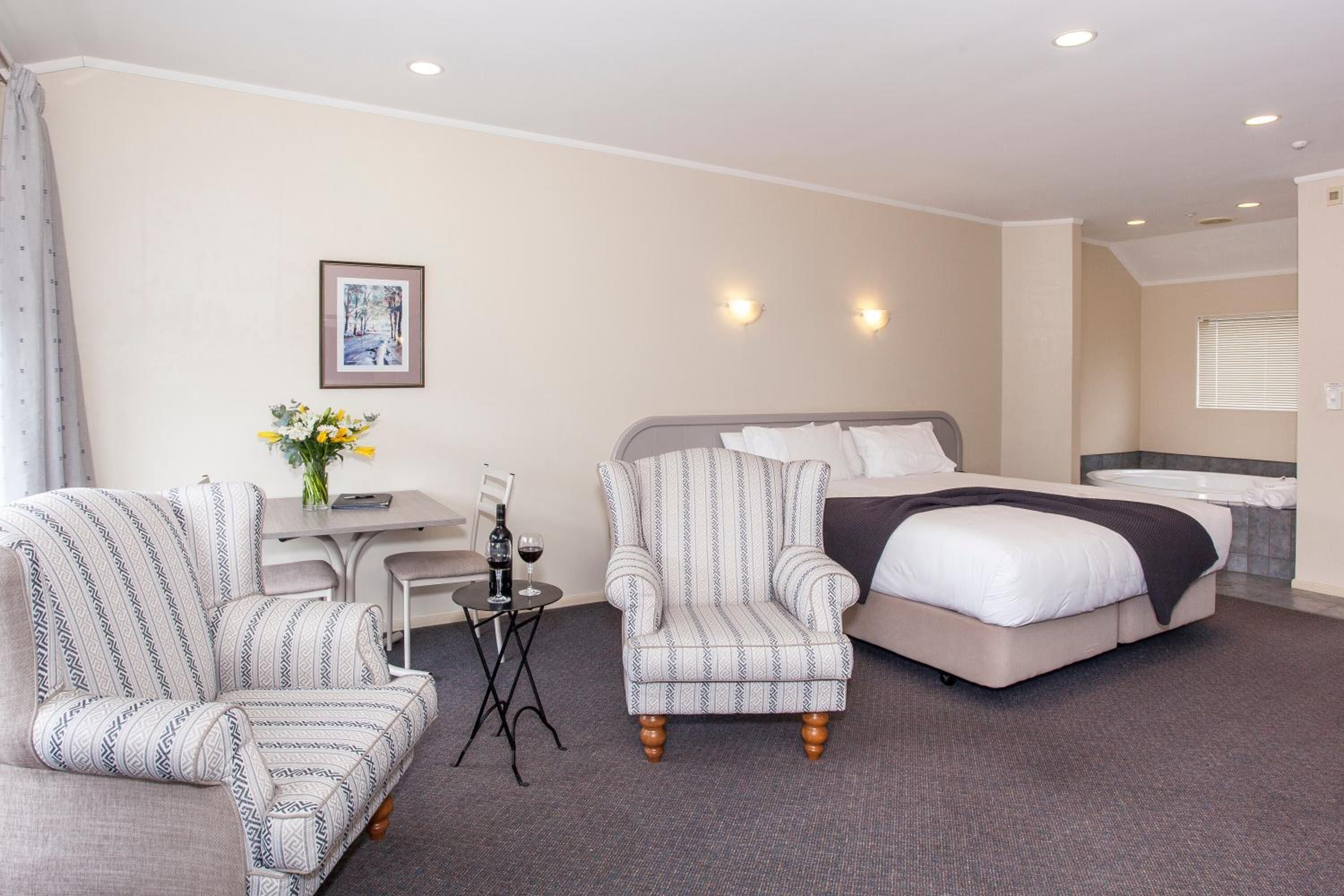 A hotel room with a bed, two armchairs, a small table with a vase of flowers, and a window with blinds.