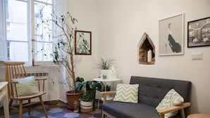 A cozy living room with a grey sofa, wooden chair, and a small table. There are potted plants and framed artwork on the walls.