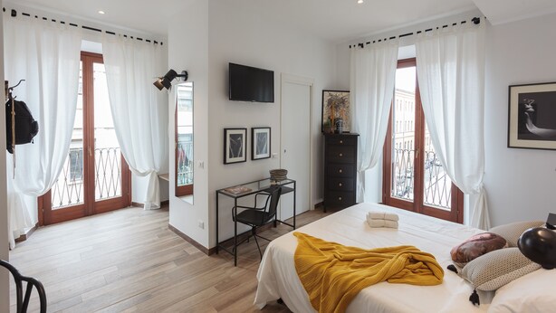 A bedroom with a bed, a desk, a chair, a TV, and a balcony with curtains.