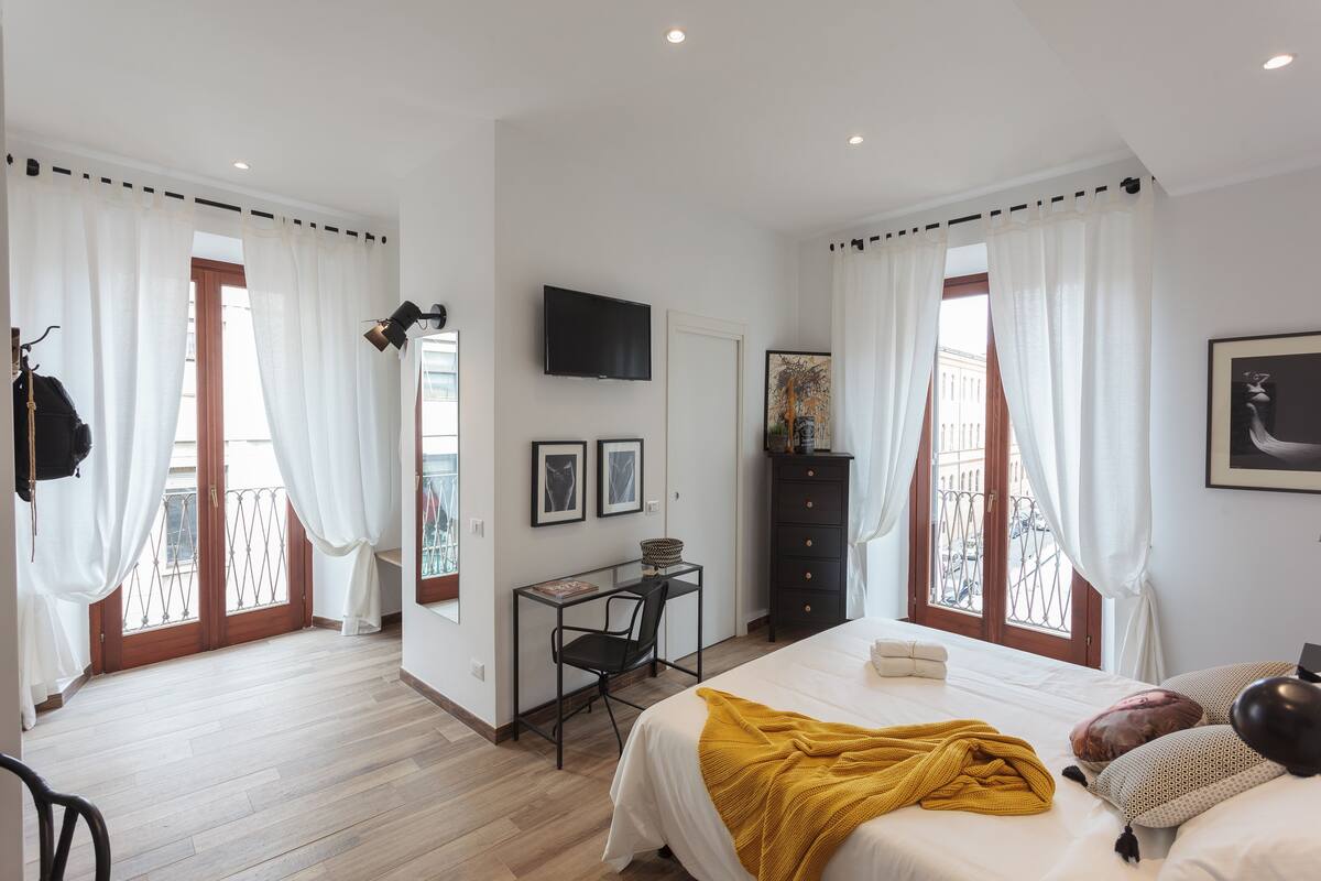 A bedroom with a bed, a desk, a chair, a TV, and a balcony with curtains.