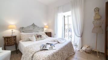 A bedroom with a large bed, two bedside tables with lamps, a dresser, a window with curtains, and a wall-mounted clock.