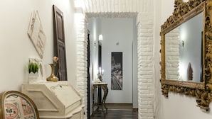 A hallway with a white brick wall, a wooden door, a mirror with an ornate frame, and a small table with a lamp.