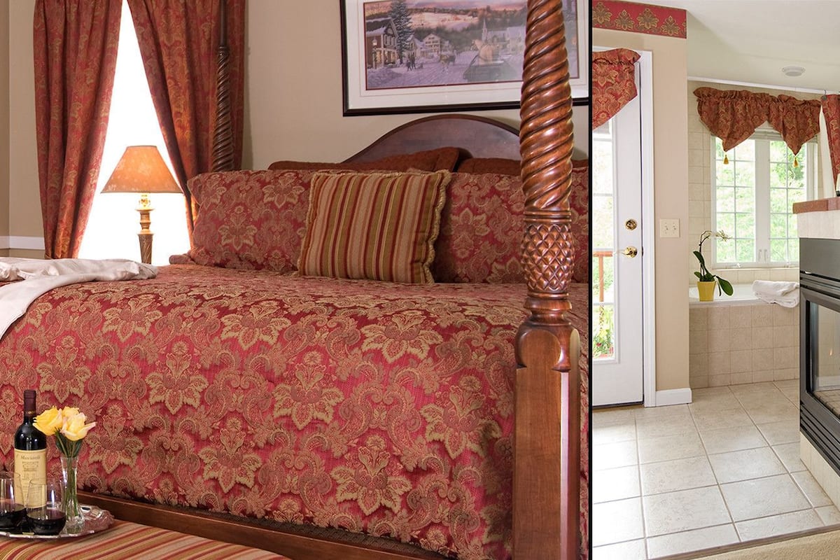 A four-poster bed with a red and gold patterned bedspread, a bedside table with a bottle and glasses, and a view of a fireplace and a window with curtains.