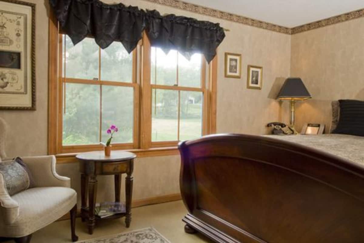 A bedroom with a large bed, a chair, a small table with a vase, and a window with curtains.