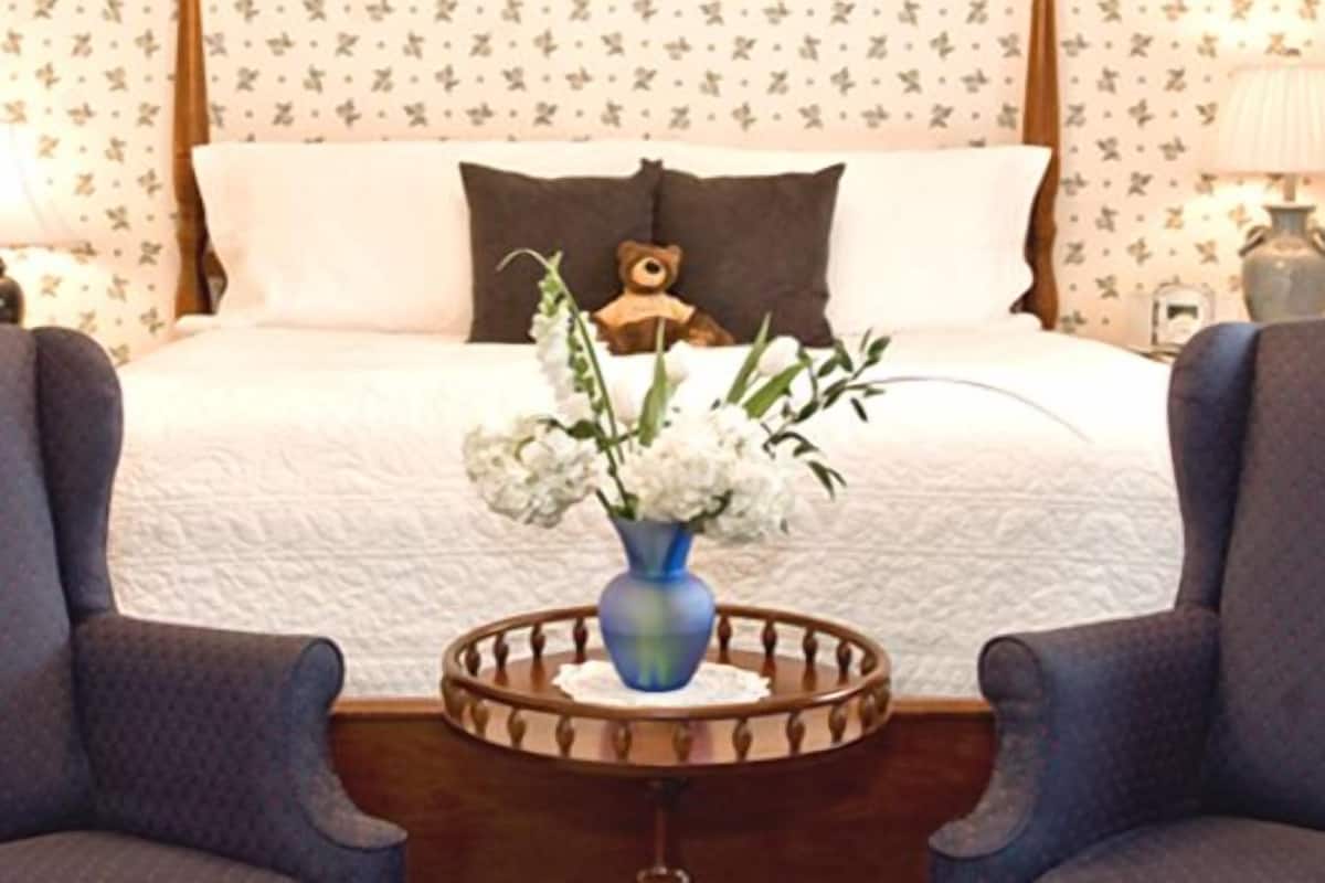 A bedroom with a bed, two armchairs, a bedside table with a vase of flowers, and a teddy bear on the bed.