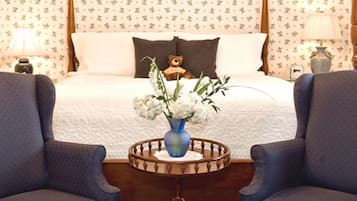 A bedroom with a bed, two armchairs, a bedside table with a vase of flowers, and a teddy bear on the bed.