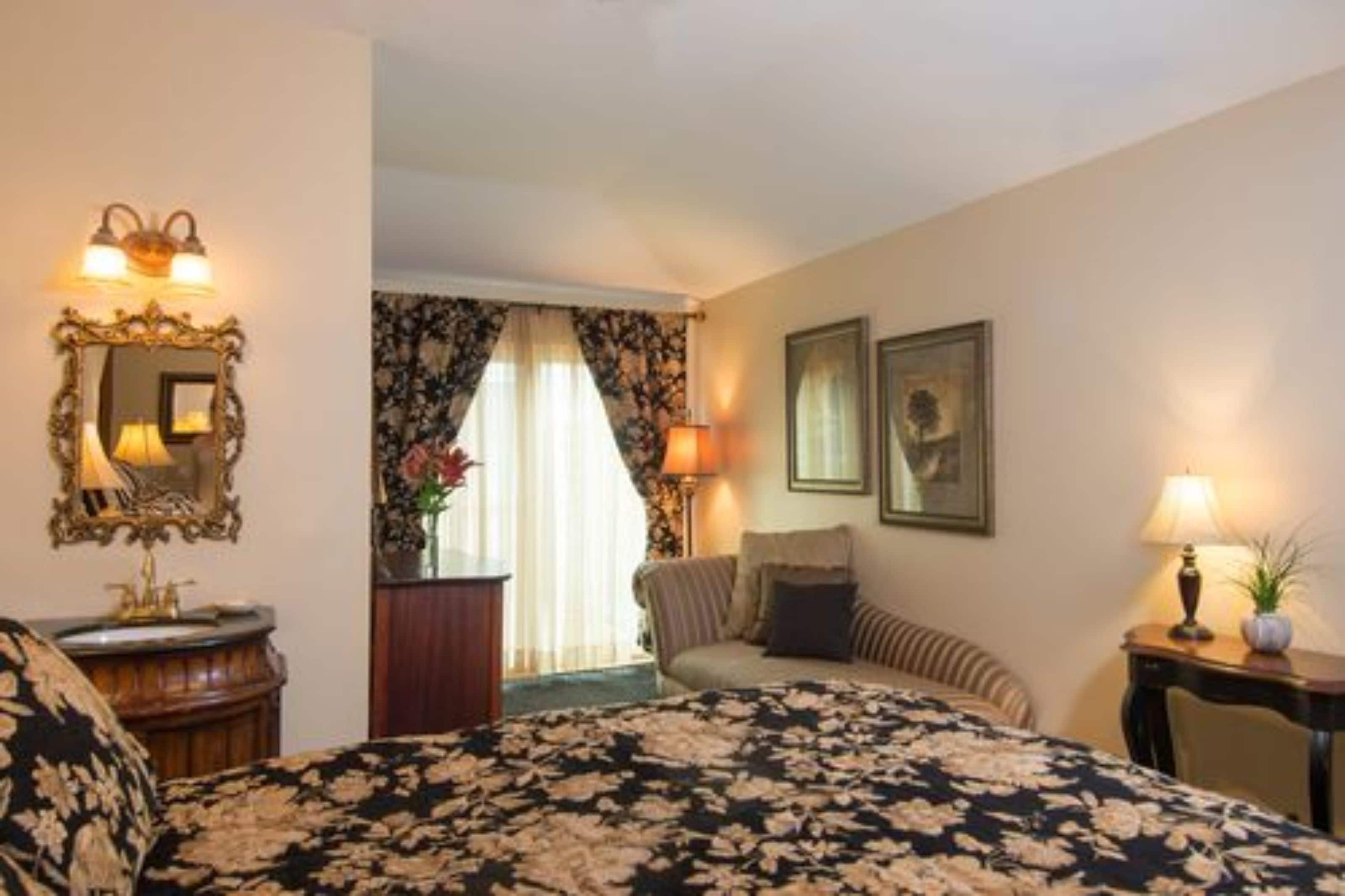A hotel room with a large bed, a vanity with a mirror, two wall-mounted lamps, a framed picture, and a window with curtains.