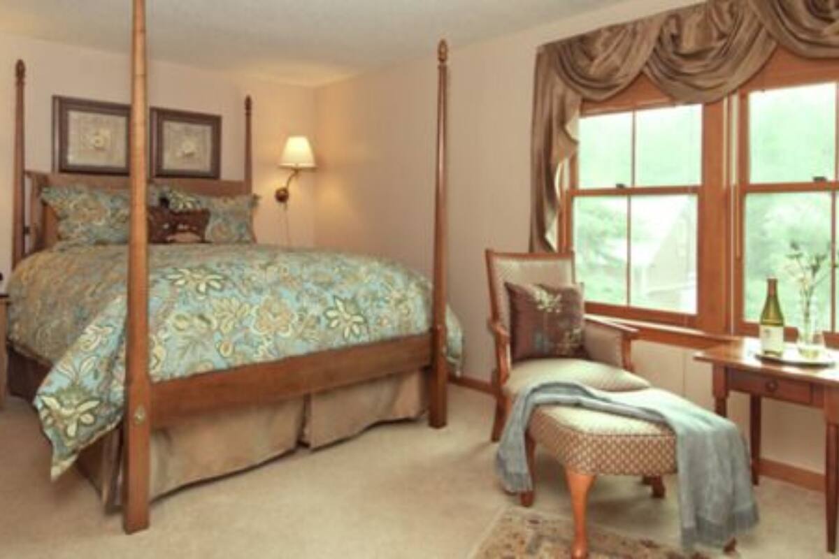 A four-poster bed with a floral bedspread, a nightstand with a lamp, a chair with a cushion, and a window with curtains.
