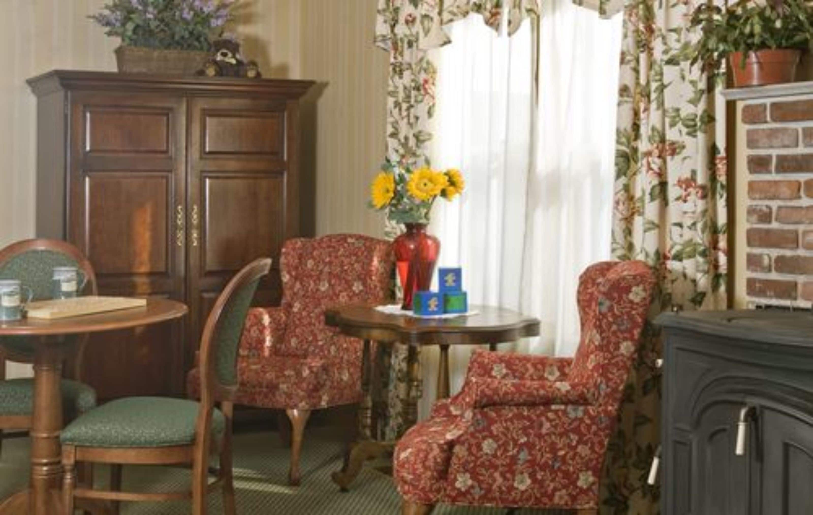 A room with a wooden cabinet, two floral-patterned chairs, a small round table with a vase of yellow flowers, and a brick-patterned fireplace.