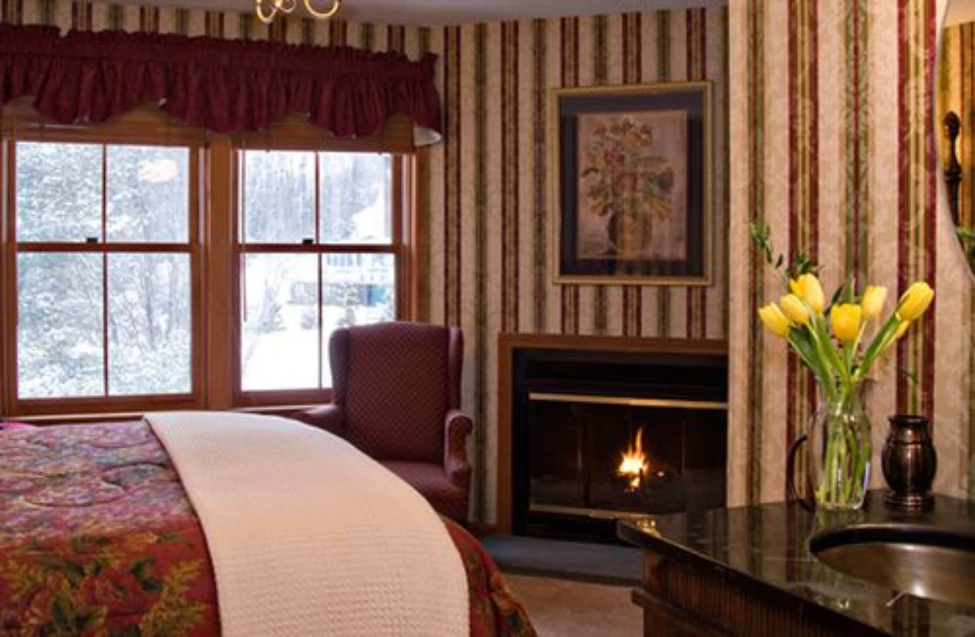 A room with a large bed, a fireplace, a window with a view of trees, and a vase with yellow flowers on a table.
