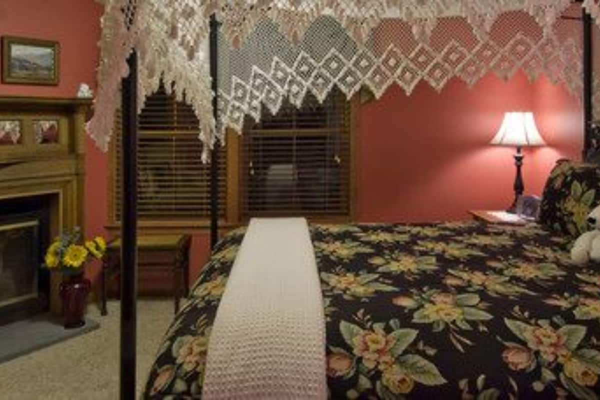 A bedroom with a fireplace, floral bedspread, lace canopy, and a nightstand with a lamp.