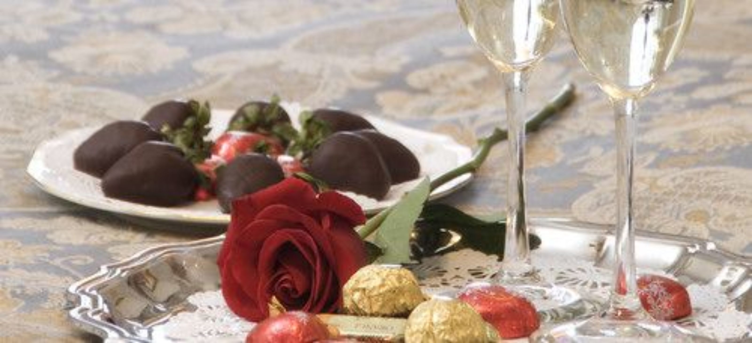 A plate of chocolate-covered strawberries, a red rose, and two glasses of white wine on a table with a lace doily.