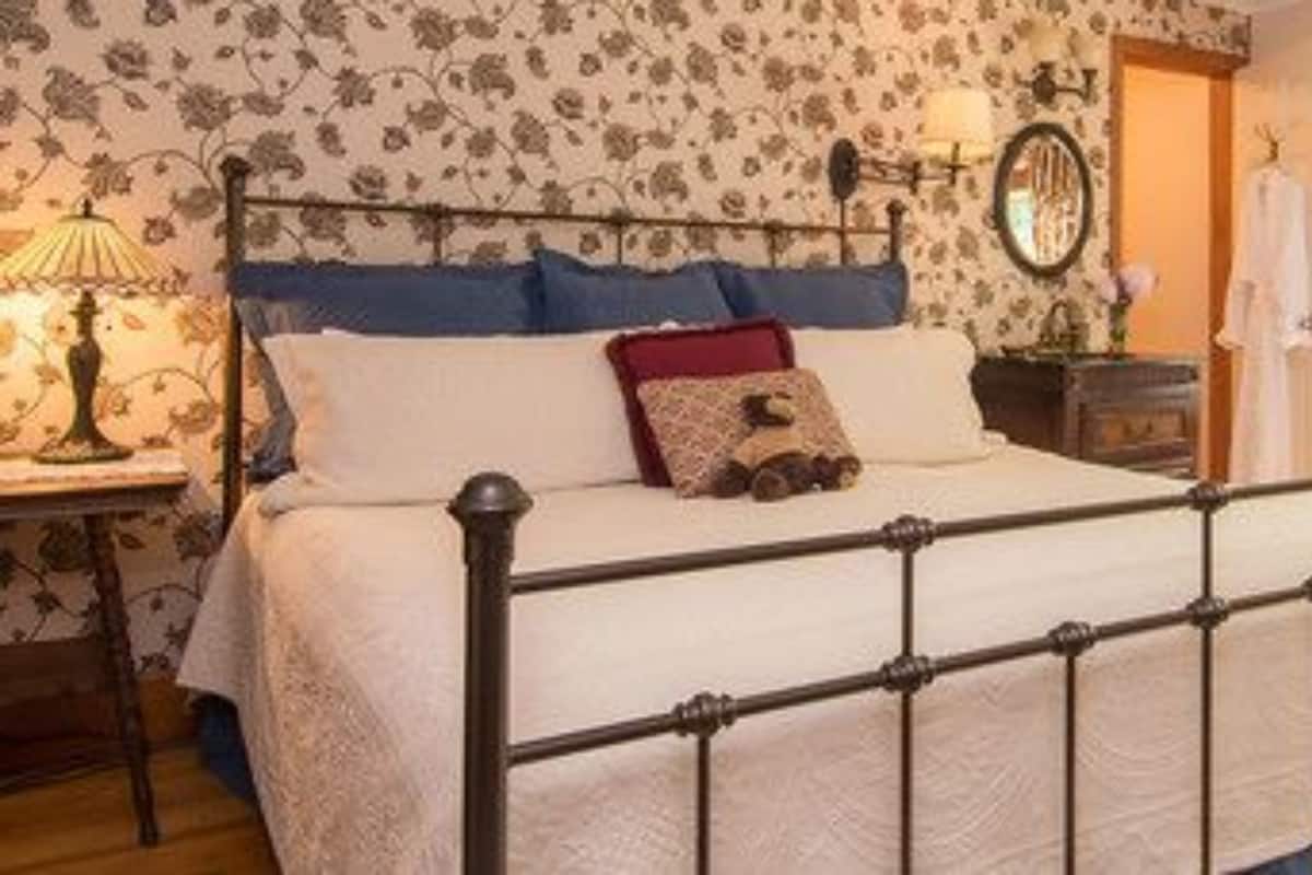 A bedroom with a metal bed frame, floral wallpaper, a bedside table with a lamp, and a window with curtains.