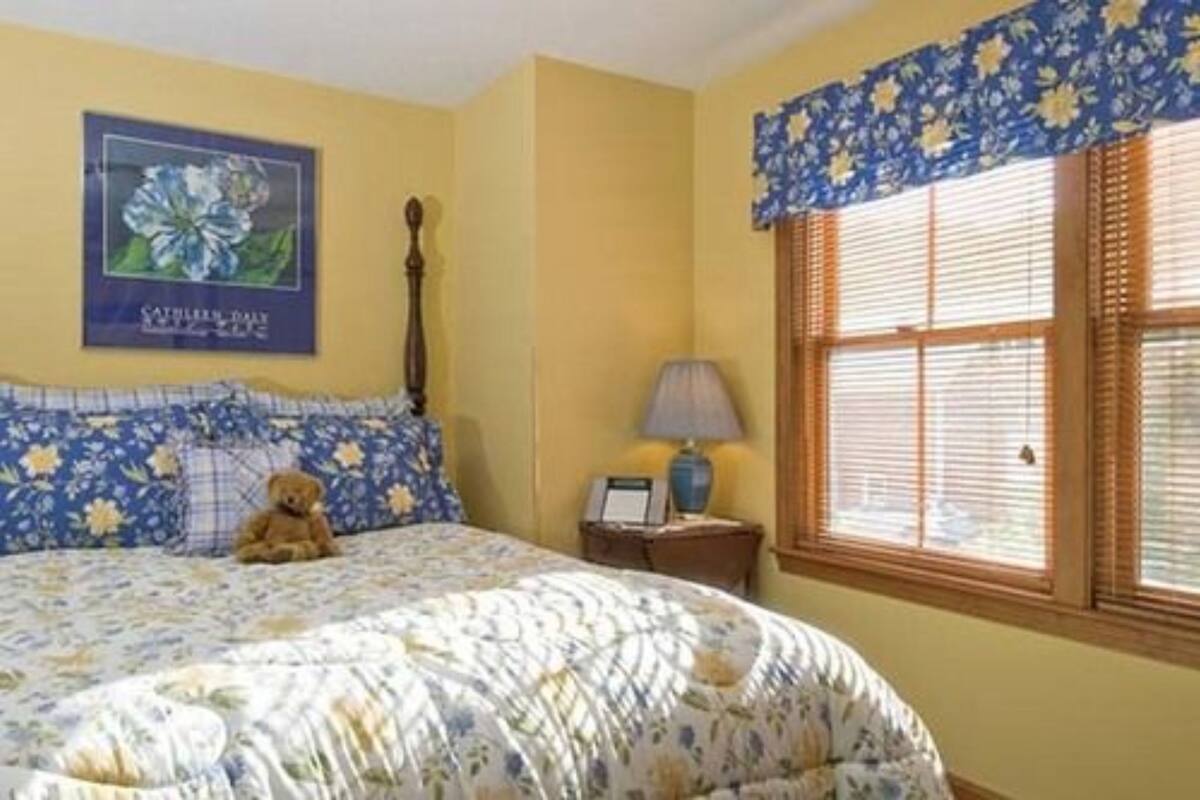 A bedroom with a floral bedspread, a teddy bear on the bed, a nightstand with a lamp, and a window with blinds.