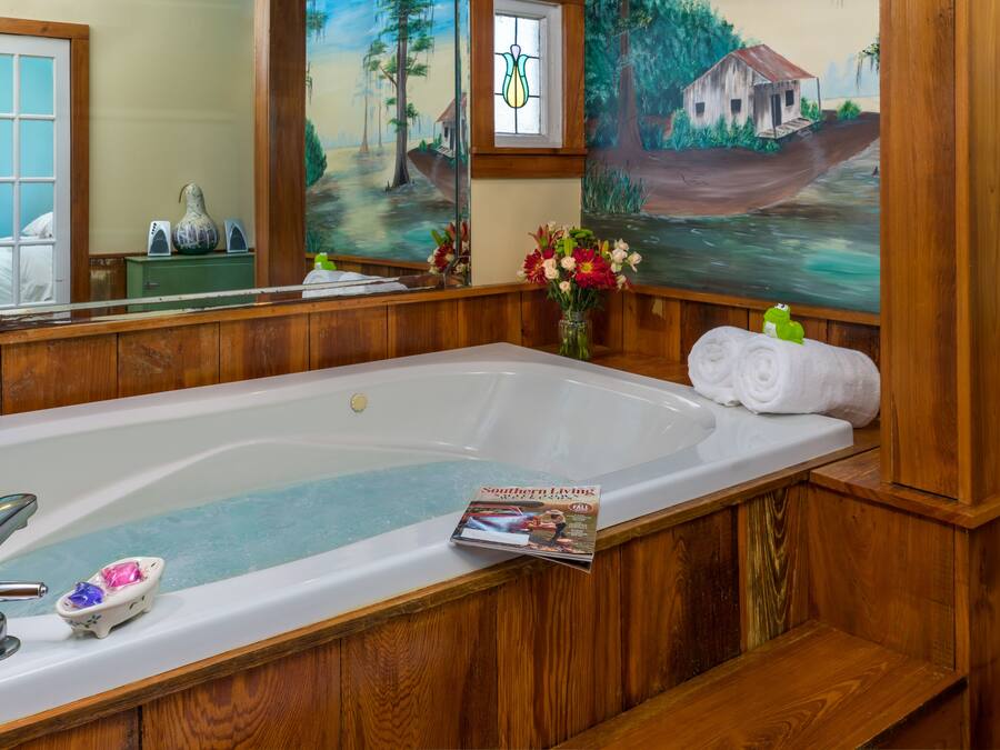 A bathroom with a large bathtub, wooden paneling, a mirror, and a painting of a rural scene.