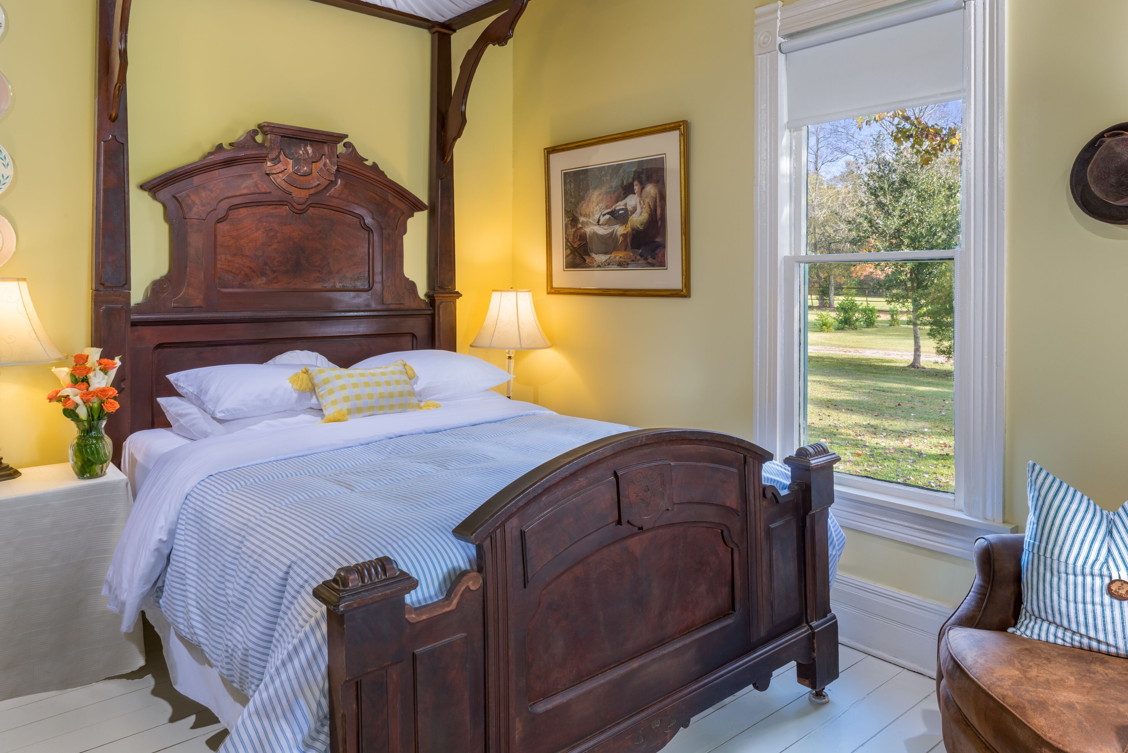 A bedroom with a large bed, a window with a view of the outdoors, a painting on the wall, and a vase with flowers on the nightstand.