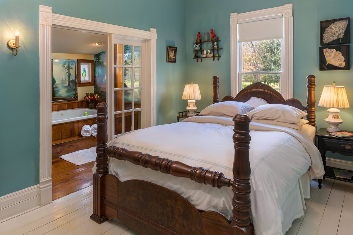 A bedroom with a large bed, a bathroom with a bathtub, and a view of trees through the window.