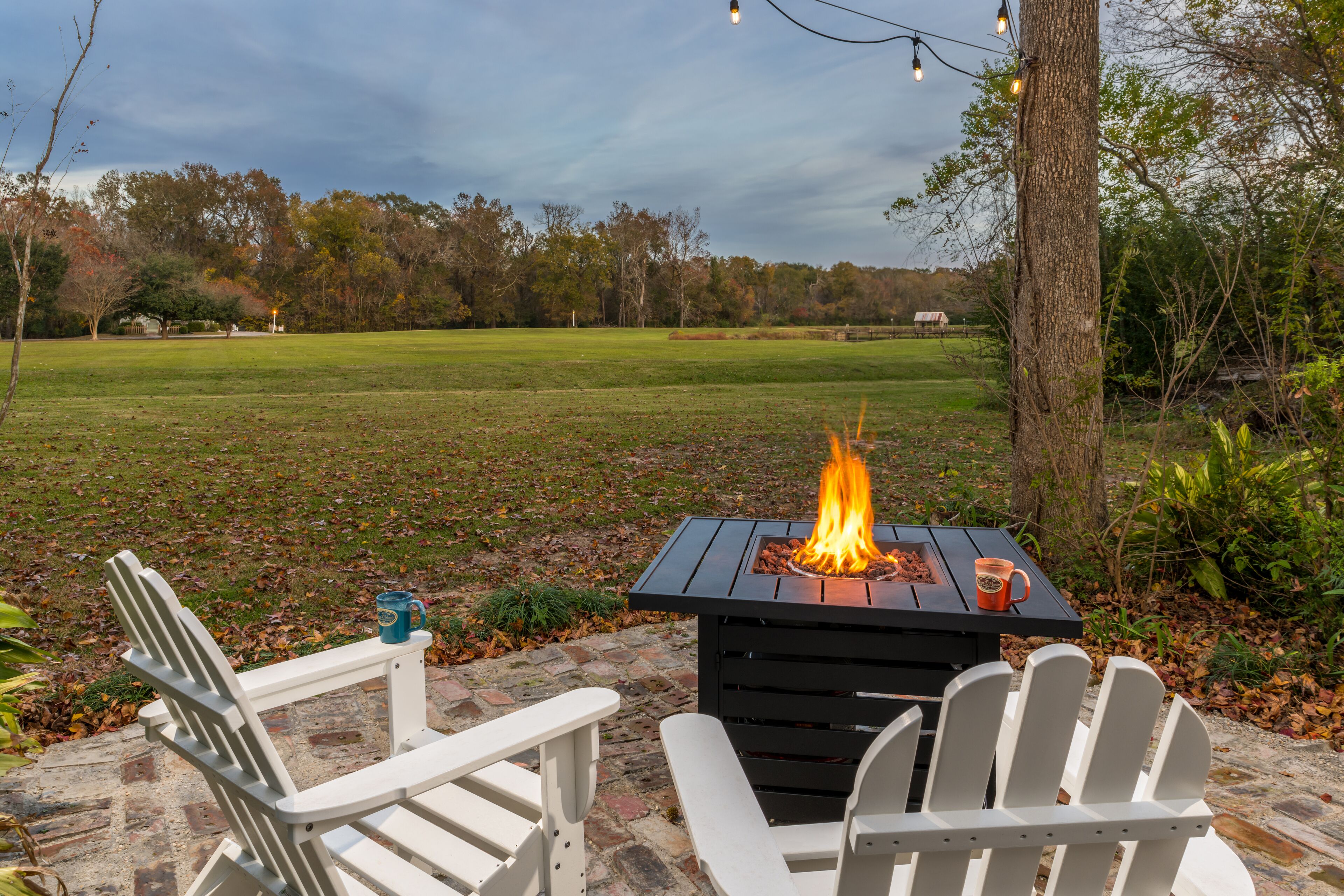 A patio with two Adirondack chairs, a small table with a fire pit, and a view of a grassy field.