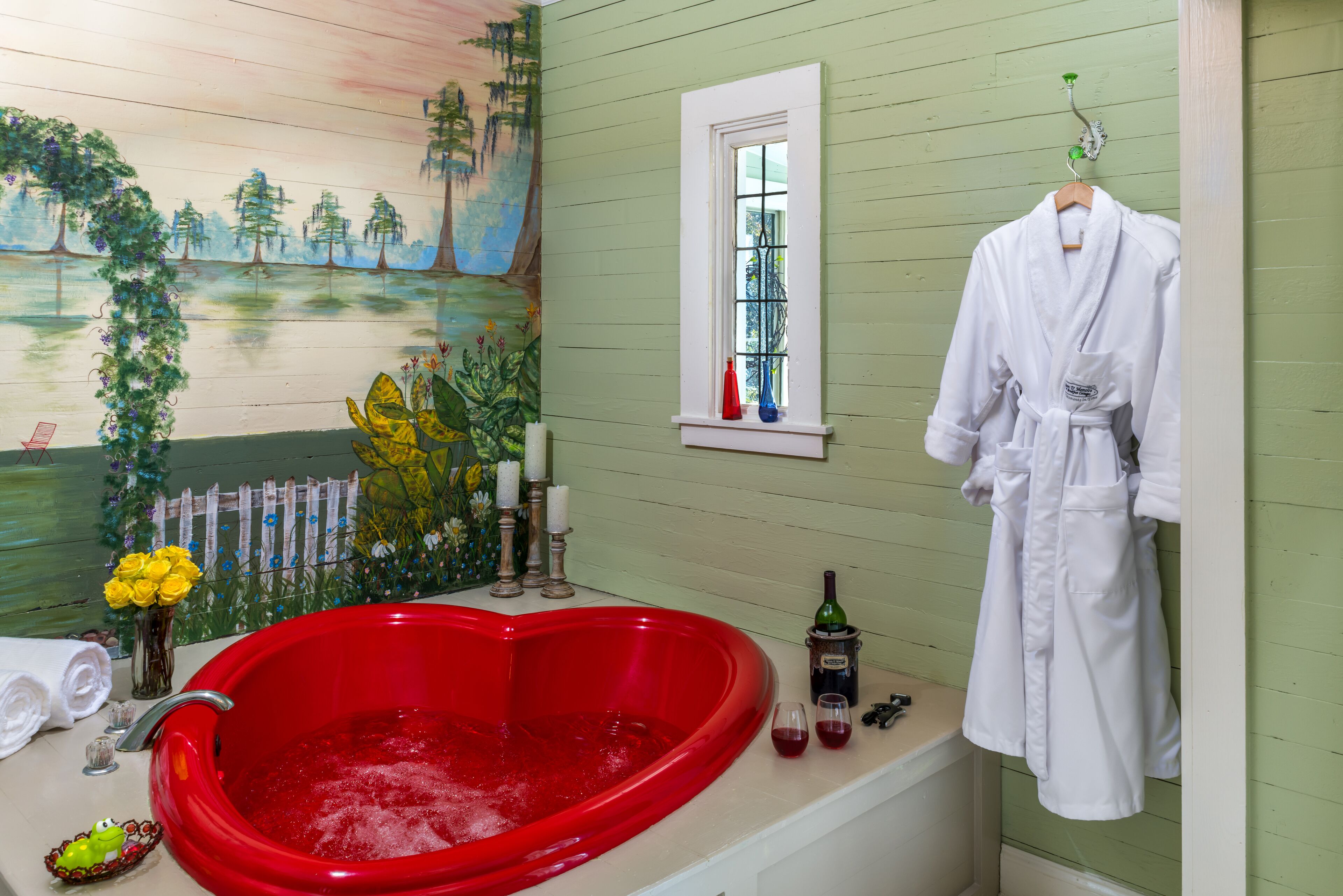 A bathroom with a heart-shaped red bathtub, a white robe hanging on the wall, and a mural of a forest scene.
