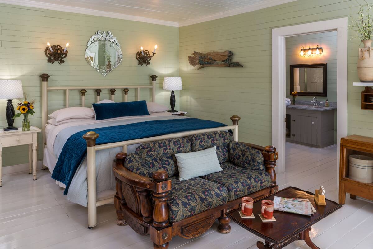 A bedroom with a bed, a sofa, a nightstand, a lamp, a mirror, and a bathroom visible through an open door.