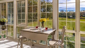 Quarto iluminado pelo sol com uma mesa, duas cadeiras, um vaso com flores e vista para uma paisagem verde através de grandes janelas.