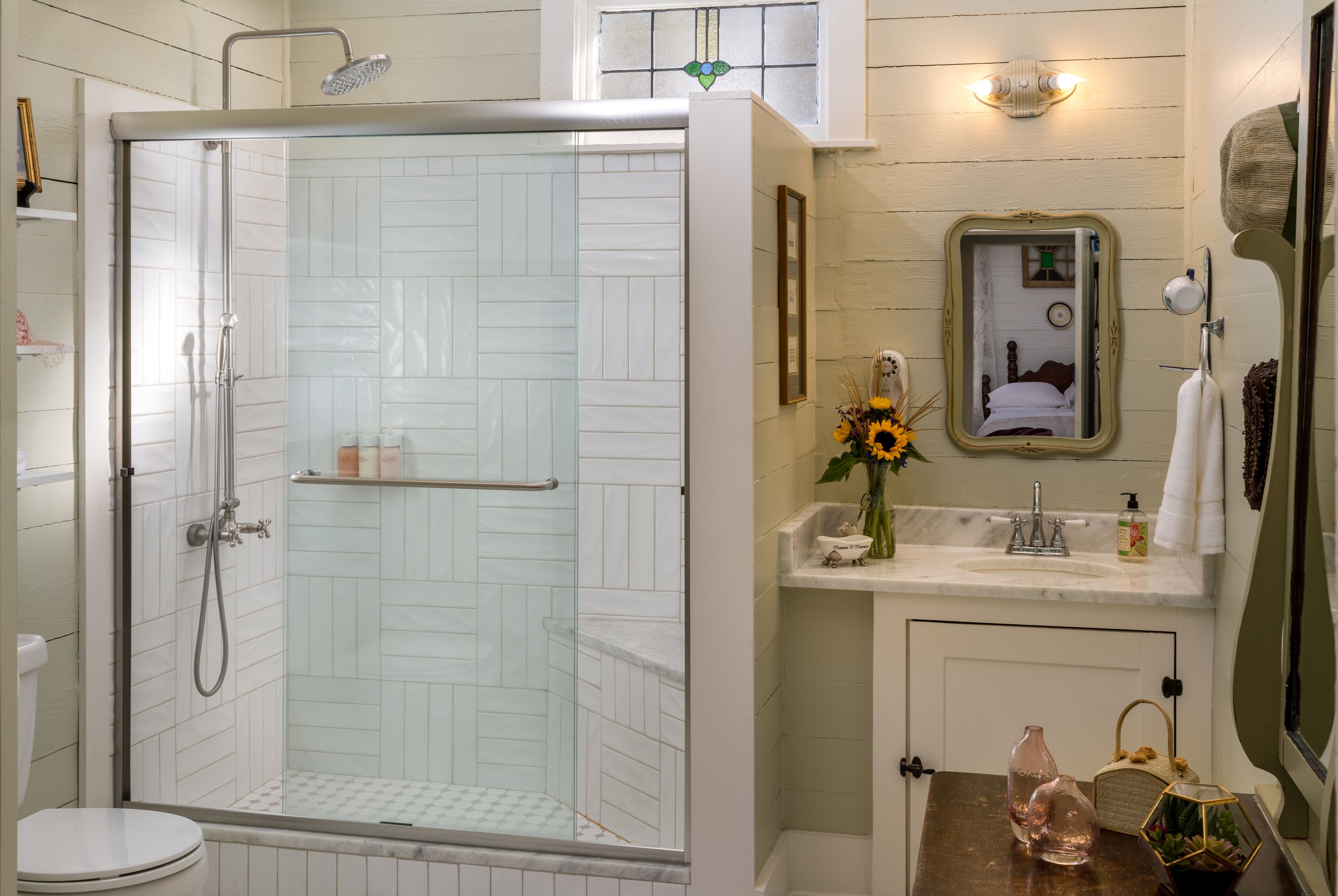 A bathroom with a glass-enclosed shower, a white toilet, a sink with a mirror, and a small shelf with toiletries.