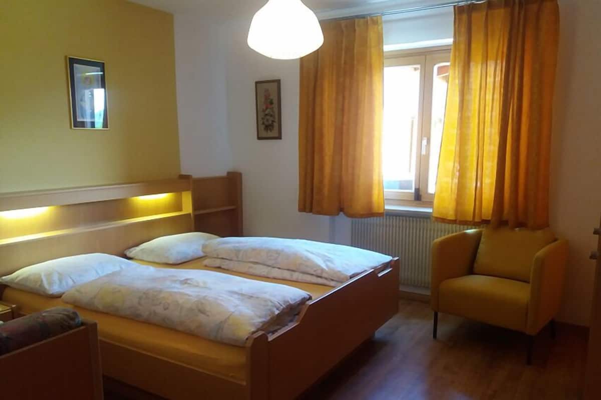 A bedroom with two beds, a chair, a window with curtains, and a radiator.