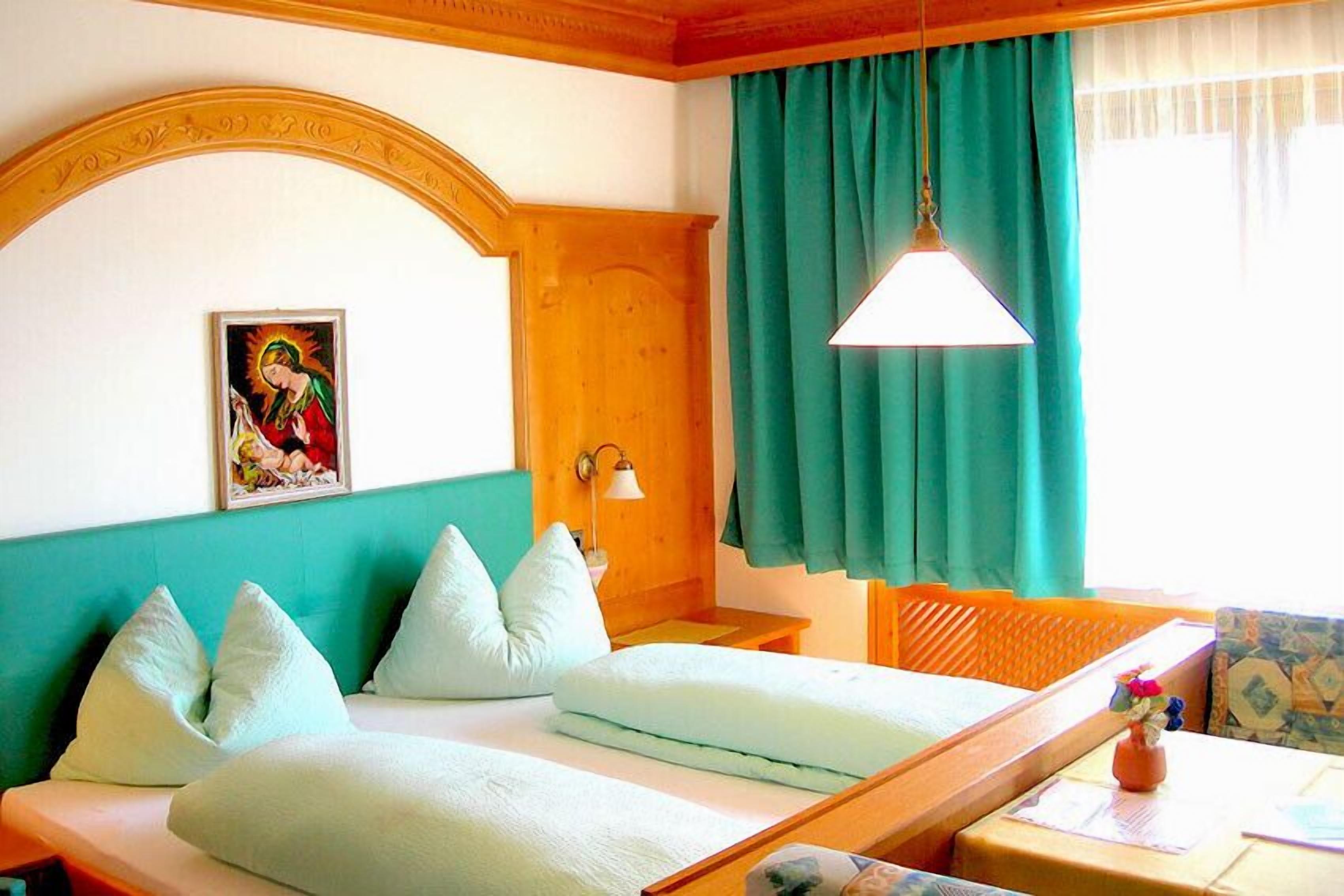 Une chambre d’hôtel avec un lit, une tête de lit en bois, un tableau, une fenêtre avec des rideaux et une table sur laquelle est posé un vase.